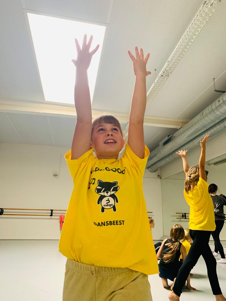 Een jong meisje in een geel T-shirt met opgeheven armen staat lachend in een felverlichte dansstudio. Andere kinderen in bijpassende shirts dansen op de achtergrond. De ruimte heeft een groot raam, spiegels, balletstangen en zichtbare plafondbuizen.