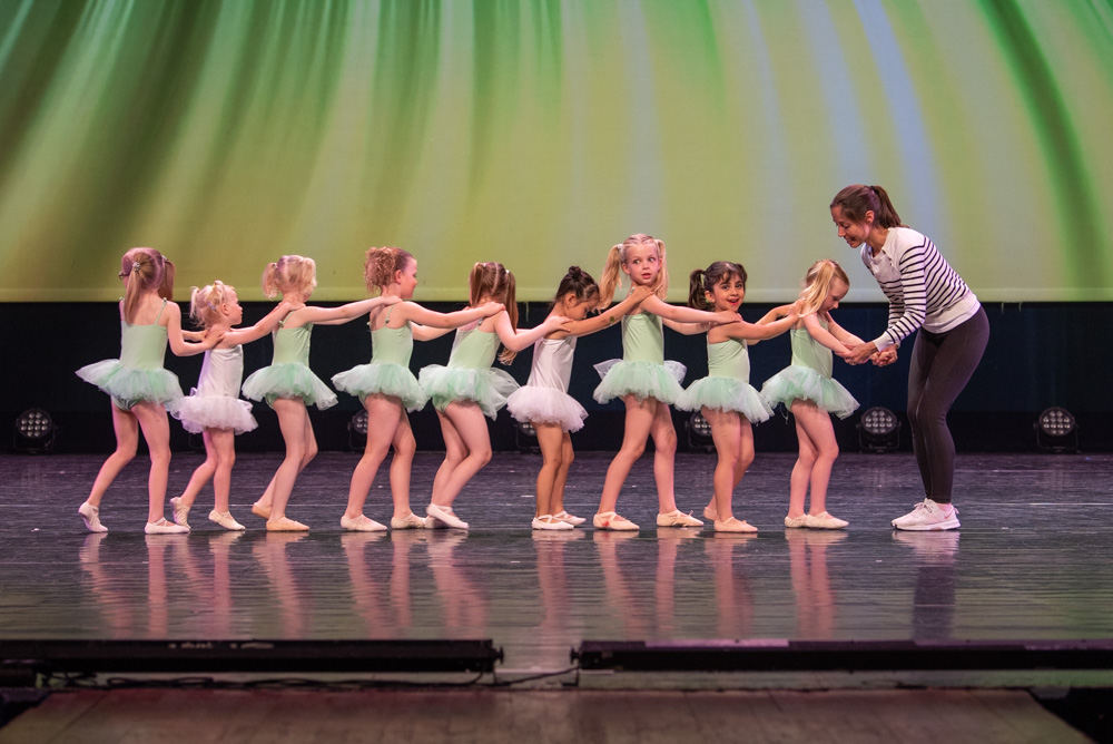 Kleuterdans 8 Een groep jonge meisjes in mintgroene tutu's staat in een rij op het podium en houdt elkaars schouders vast. Een volwassene, casual gekleed in een zwarte broek en een gestreepte top, staat tegenover hen en houdt de hand van het eerste meisje vast. De podiumlampen werpen een zachtgroene gloed op de achtergrond.