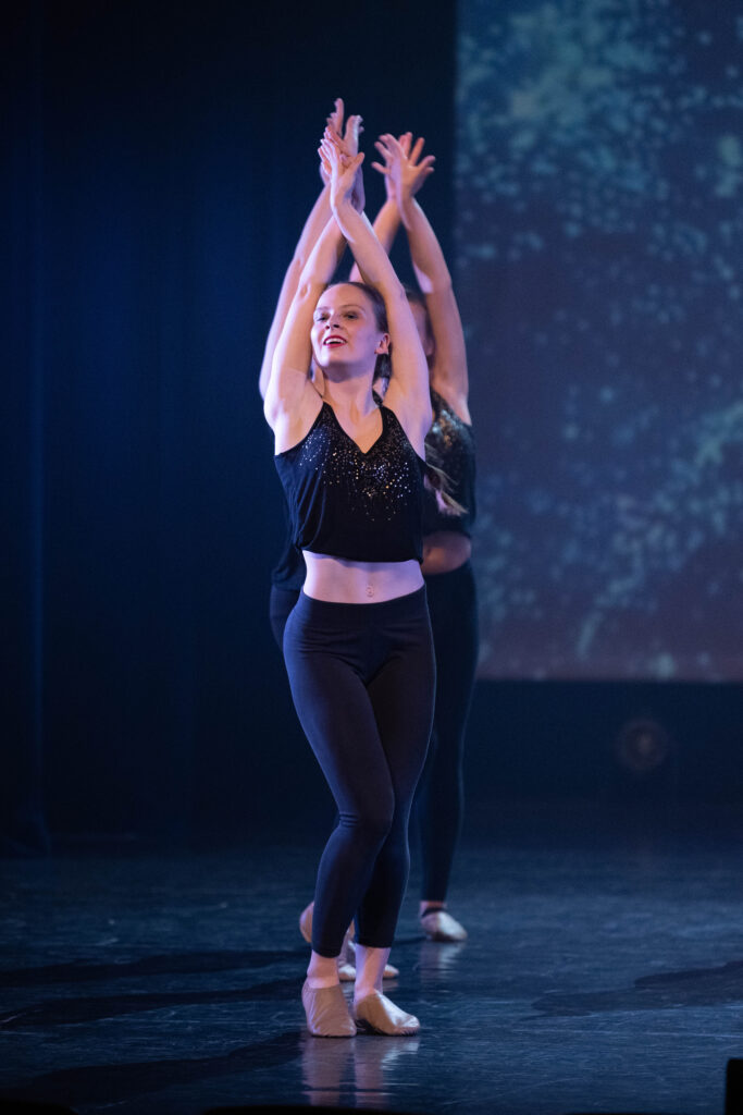Twee dansers staan in een rij op het podium van CC De Spil in Roeselare, met geheven armen en handen die elkaar boven hun hoofd raken. De danseres op het podium, glimlachend, draagt een glinsterend zwart mouwloos topje – een treffende weergave van de levendige geest van jazzdansen.
