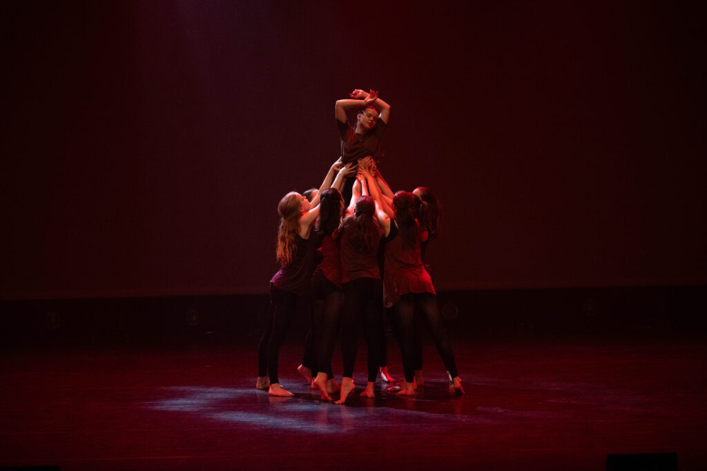 Hedendaagse dans 2 Vijf dansers in donkere kleding tillen een zesde danseres boven zich op een schemerig podium. De geheven danseres poseert met haar armen omhoog en gebogen, terwijl de anderen haar ondersteunen en een dynamische, expressieve vorm vormen tegen een donkerrode achtergrond.