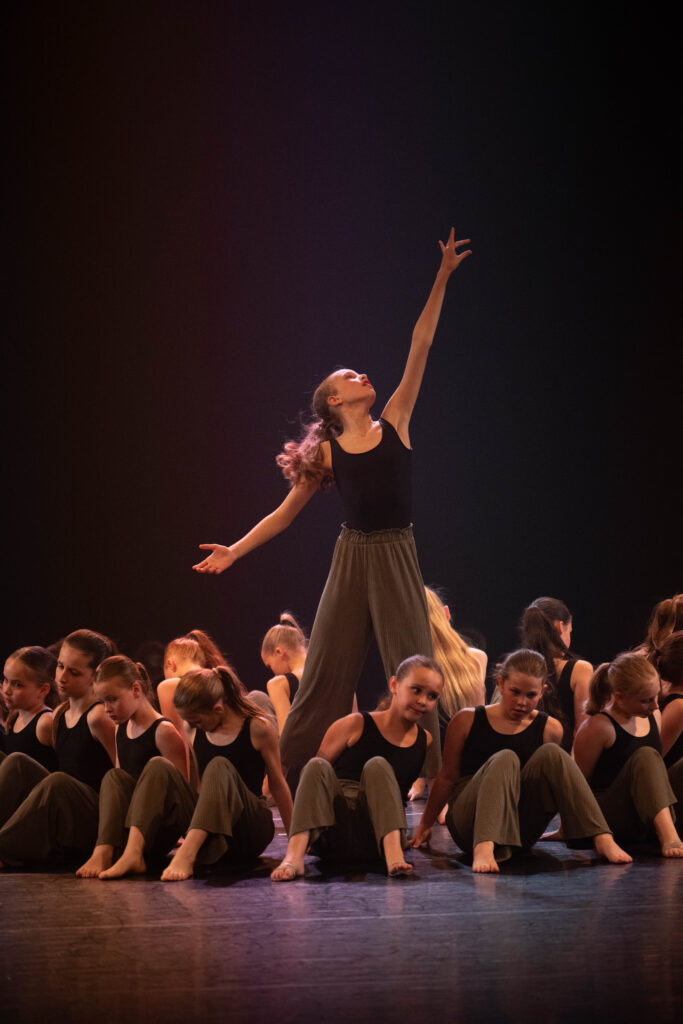 Hedendaagse dans 1 Een groep dansers op blote voeten zit op het podium in bijpassende zwarte tops en losse olijfgroene broeken. Eén danser staat rechtop in het midden en reikt met één arm omhoog, verlicht door het podiumlicht, wat een dramatisch middelpunt vormt tegen de donkere achtergrond.