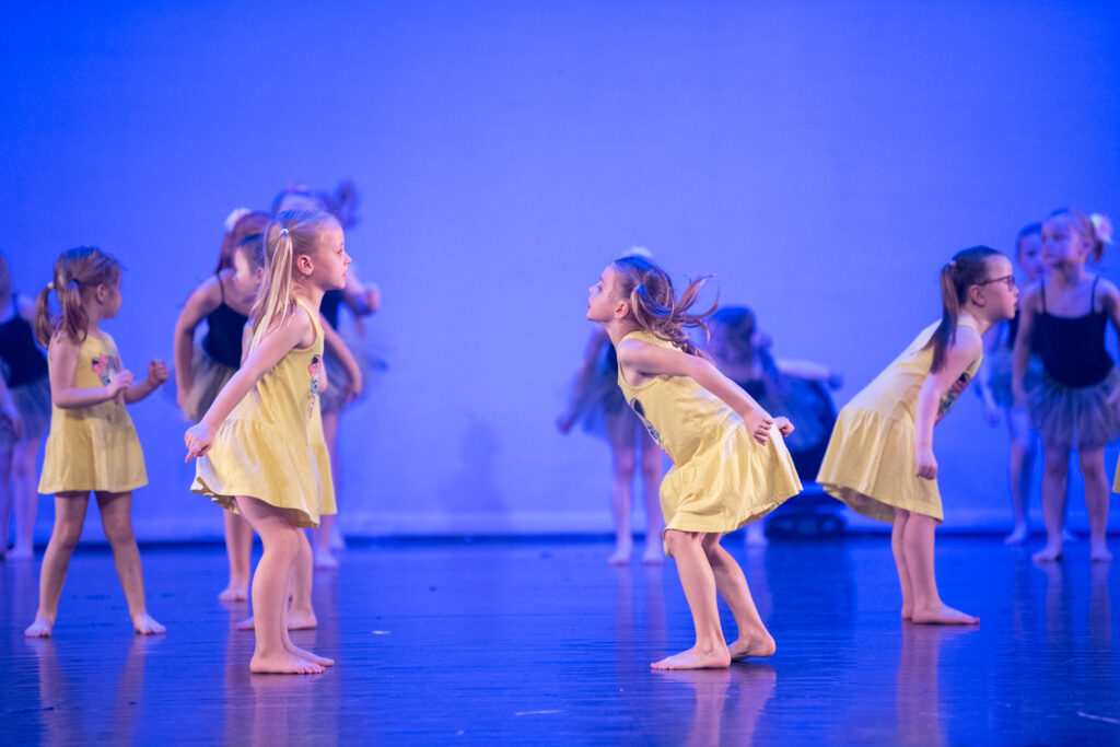Een groep jonge kinderen treedt op op het podium, voornamelijk meisjes in gele jurken, dansend in gecoördineerde poses. De achtergrond is blauw verlicht en de meisjes lijken betrokken en expressief, sommigen leunen voorover en anderen staan rechtop.