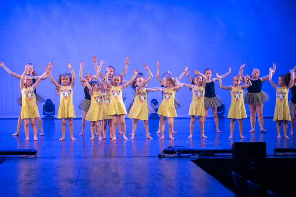 Kleuterdans 6 Een groep jonge kinderen danst energiek op het podium. De meesten dragen gele jurken met dierengezichten, terwijl een paar achterin grijze tutu's dragen. Ze lopen op blote voeten, met hun armen omhoog, tegen een blauw verlichte achtergrond, en voeren een gecoördineerde routine uit.