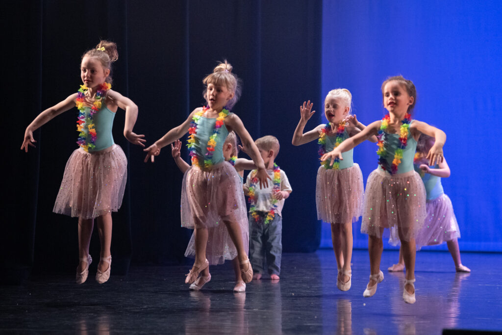 Een groep jonge kinderen in glinsterende roze tutu's, kleurrijke bloemenkransen en balletschoentjes treedt op op het podium. Een aantal meisjes is midden in een sprong, terwijl anderen erachter staan, tegen een blauw-zwarte achtergrond.