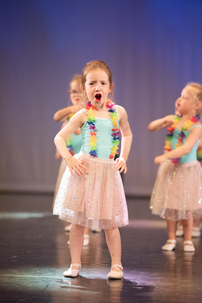 Een jong meisje in een lichtblauw turnpakje en een glinsterende rok treedt op het podium op, gekleed in een kleurrijke bloemenkrans. Haar mond staat open alsof ze zingt of schreeuwt, en andere kinderen in soortgelijke kleding dansen op de achtergrond tegen een effen achtergrond.