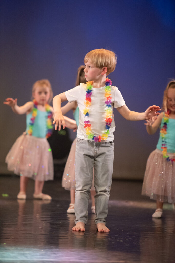 Een jonge jongen in een wit T-shirt, spijkerbroek en een kleurrijke bloemenkrans staat blootsvoets op het podium, met opgeheven armen midden in de dans. Achter hem voeren drie meisjes in pastelkleurige tutu's, blauwe topjes en bloemenkransen soortgelijke dansbewegingen uit, allemaal tegen een zacht verlichte blauwe achtergrond.