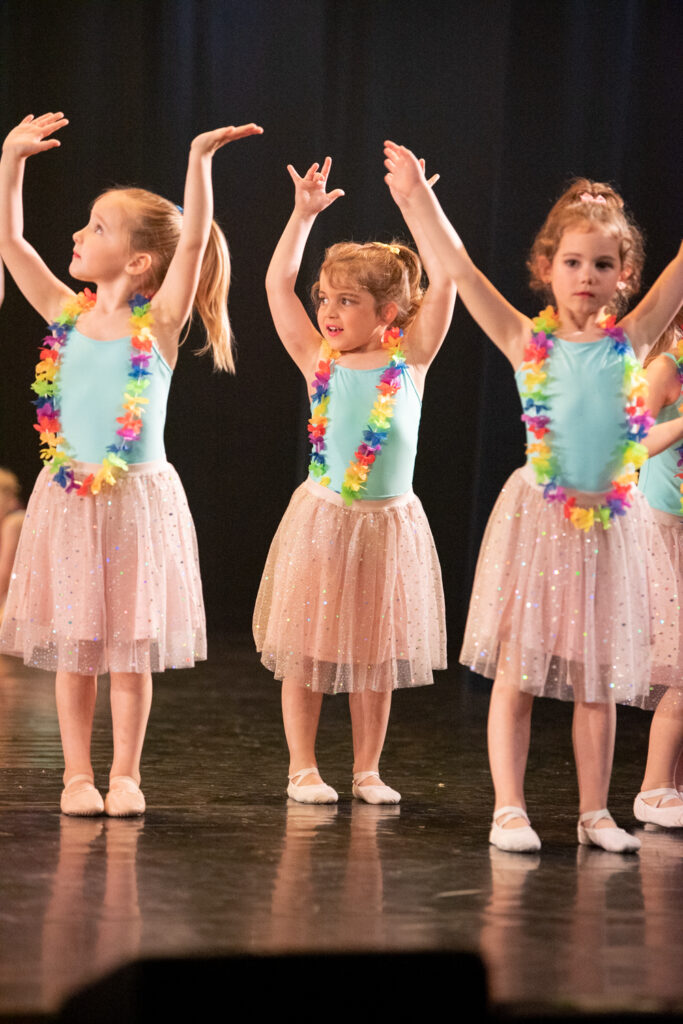 Drie jonge meisjes in blauwe maillots, glinsterende roze rokken en kleurrijke bloemenkransen staan met opgeheven armen op het podium. Ze dragen balletschoenen en voeren een dansroutine uit, met een donkere achtergrond die hun felle kostuums accentueert.
