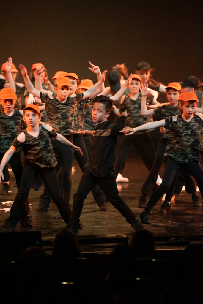 Een groep kinderen voert een dans op op het podium. De meesten dragen oranje petten, camouflageshirts en zwarte broeken, terwijl één jongen in het midden helemaal in het zwart gekleed is. Ze nemen expressieve poses aan met uitgestrekte armen. De achtergrond is donker en het publiek kijkt in de schaduw toe.