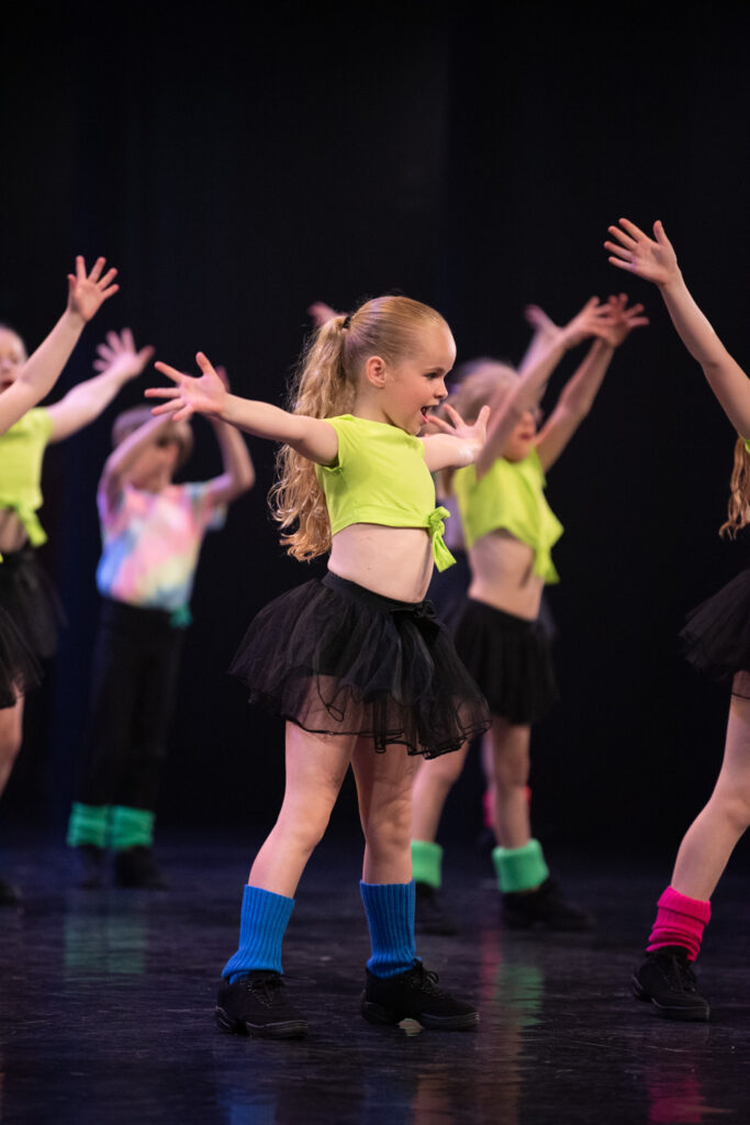 Jonge Funky Dance meisjes voeren een dansroutine uit op het podium. Het centrale meisje draagt een neongroene crop top, een zwarte tutu, zwarte schoenen en blauwe beenwarmers. Ze glimlacht met opgeheven armen. Andere kinderen in soortgelijke kleding dansen op de achtergrond tegen een donkere achtergrond.