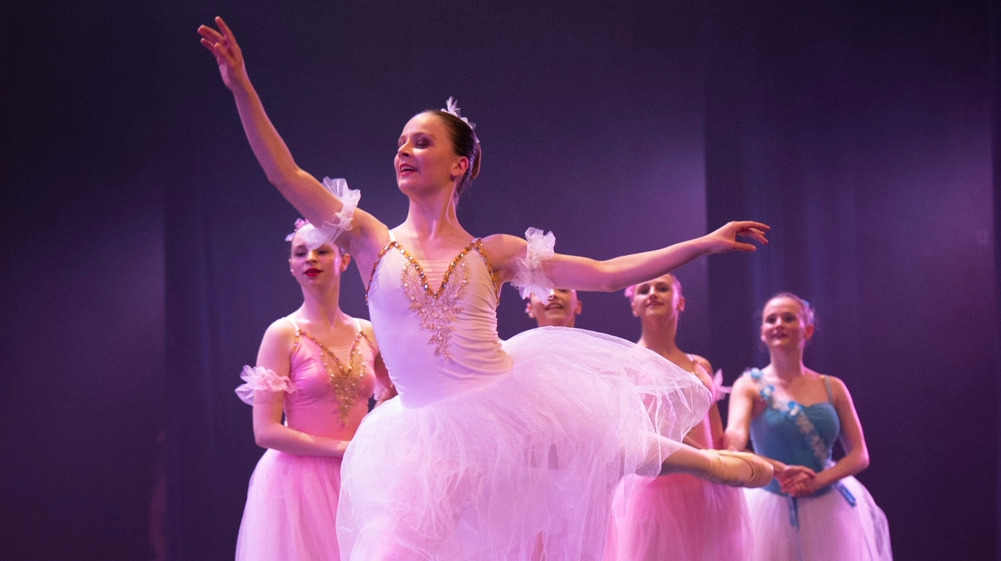 Pointes & Repertoire 1 Een ballerina in een witte tutu danst en pointe midden op het podium, omringd door pastelkleurige danseressen, die een prachtig repertoire uitvoeren onder paarse lampen.