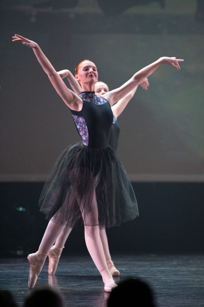 Een balletdanseres in een zwarte tutu, een maillot met geprinte zijkanten en een lichtgekleurde panty treedt op. Haar armen zijn sierlijk geheven en ze staat op haar tenen. Achter haar is een andere danseres gedeeltelijk zichtbaar, eveneens in balletkleding, tegen een donkere, zacht verlichte achtergrond.