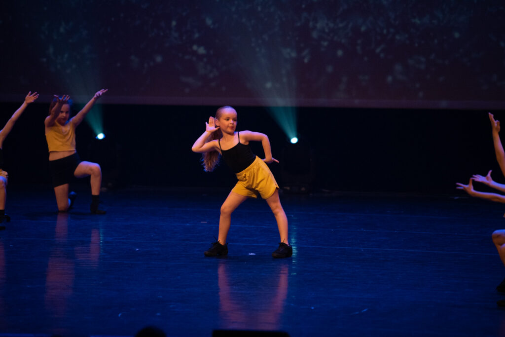 Een jong meisje in een gele korte broek en een zwart topje voert een dansroutine uit op het podium, waarbij ze een pose aanneemt met haar hand bij haar oor. Andere dansers in soortgelijke outfits staan om haar heen. Blauwe podiumlichten en een donkere sterrenachtergrond creëren een dramatische sfeer.