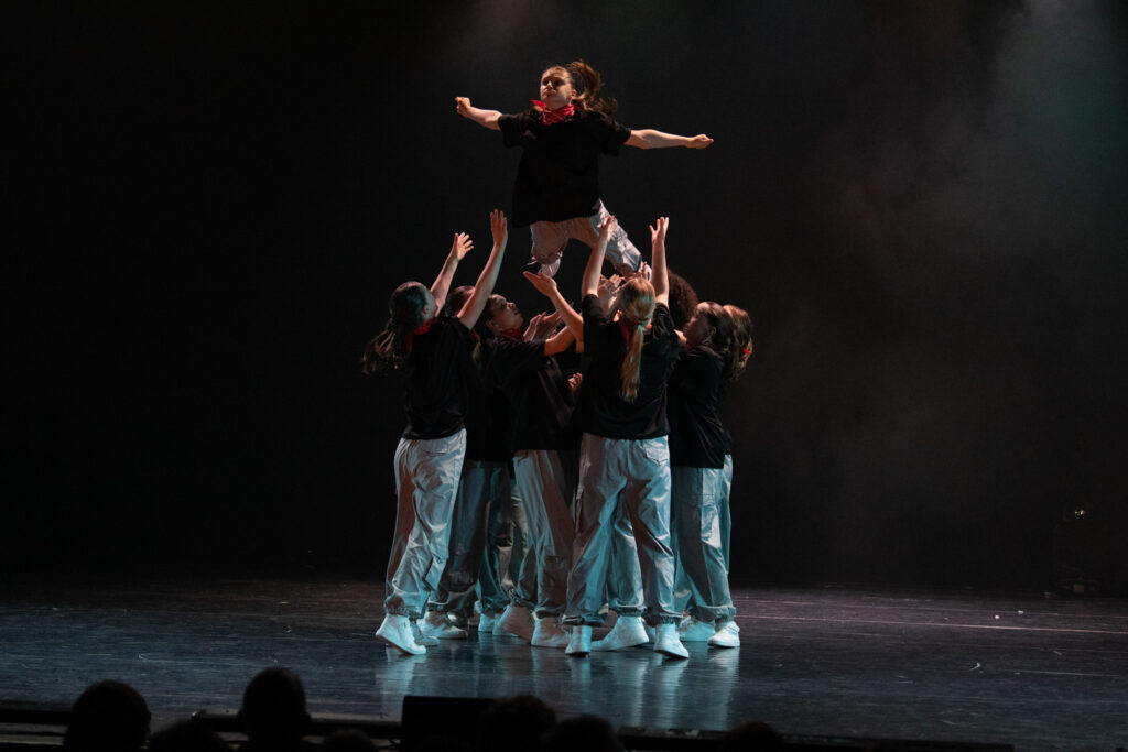 Een groep dansers in zwarte shirts en lichte broeken tilt een teamgenoot de lucht in op een schemerig podium. De danseres spreidt haar armen wijd, terwijl de anderen haar van onderaf ondersteunen. Rook of mist zorgt voor sfeer op de achtergrond.