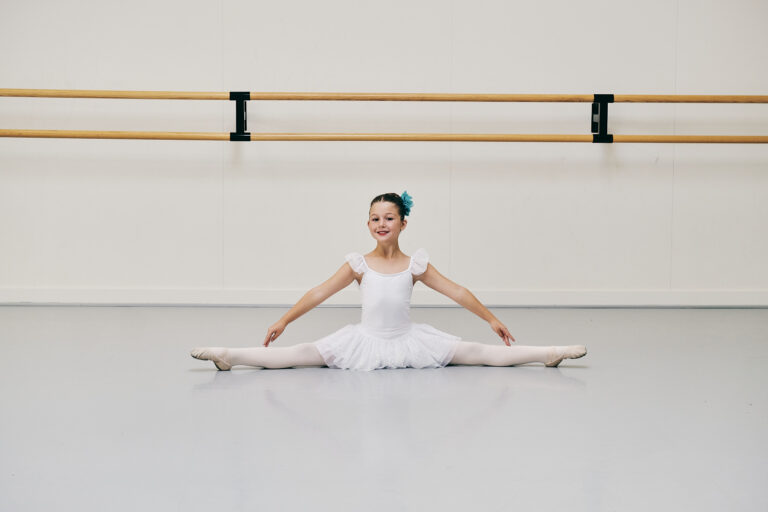 Kinderontwikkeling: Waarom dans belangrijk is voor Je Kind 1 Jong meisje in een wit balletpakje lacht terwijl ze een split doet, blauwe bloem in haar haar - kinderontwikkeling in de dansstudio.
