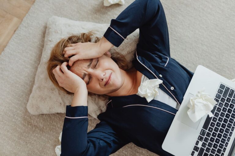 Een vrouw in pyjama ligt op een kussen en vertoont tekenen van een burn-out, gefrustreerd naast haar laptop en verfrommelde tissues op de vloer van het tapijt.